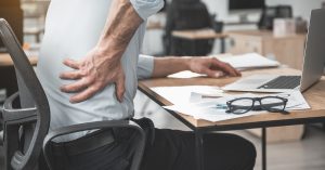 Employer having pain in back. He holding it by hand while sitting at table during job. Worker with bad state of health concept; blog: prevent back pain at work
