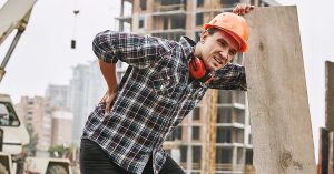 Hard work. Construction worker in protective helmet feeling back pain while working at construction site. Building construction. Pain concept. Dangerous job; blog: Top 10 Jobs That Cause Back Pain